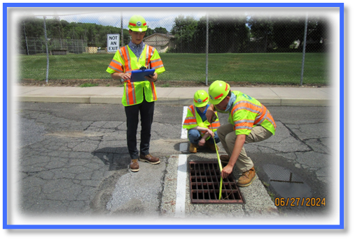 "Three individuals wearing safety vests and hard hats are inspecting a storm drain at the edge of a paved road. Two individuals are crouched beside the drain, using a measuring tape across the metal grate. Another individual stands nearby holding a clipboard. A chain‑link fence, grass, and a sign reading ‘Not an Exit’ are visible in the background. The image is dated 06/27/2024 in the lower right corner."
